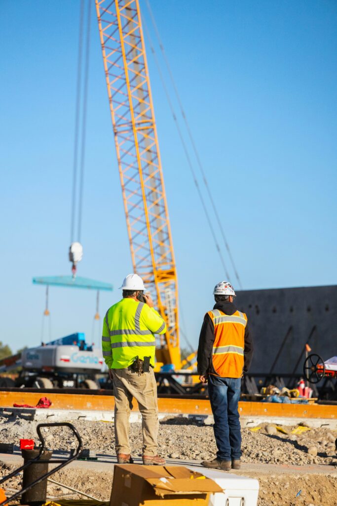 Two construction workers wearing hard hats and high visibility safety vests are standing next to each other overseeing a job site. The man on the left is on the phone. There is a construction crane in the background.